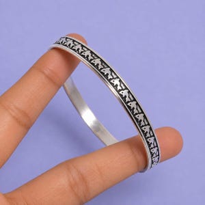 Könnte beinhalten: Ein silberfarbener Metallarmreif mit einem durchgehenden eingravierten Muster aus kleinen Fischen auf der Außenseite. Das Armband hat eine glatte, polierte Innenseite und ein dunkles, vertieftes Design, das das Fischmotiv hervorhebt.