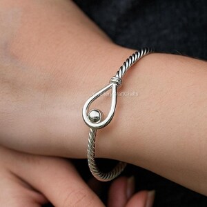 Könnte beinhalten: Ein silberfarbenes Armreif-Armband mit einem gedrehten Seildesign. Das Armband hat ein tropfenförmiges Mittelstück mit einem kleinen, runden Akzent. Der Schmuck wird am Handgelenk getragen.