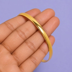 Könnte beinhalten: Ein glatter, polierter goldfarbener Armreif mit einem abgerundeten, durchgehenden kreisförmigen Band. Die Metalloberfläche weist ein reflektierendes Hochglanz-Finish auf, das das Licht einfängt und ein helles, gleichmäßiges Erscheinungsbild erzeugt.