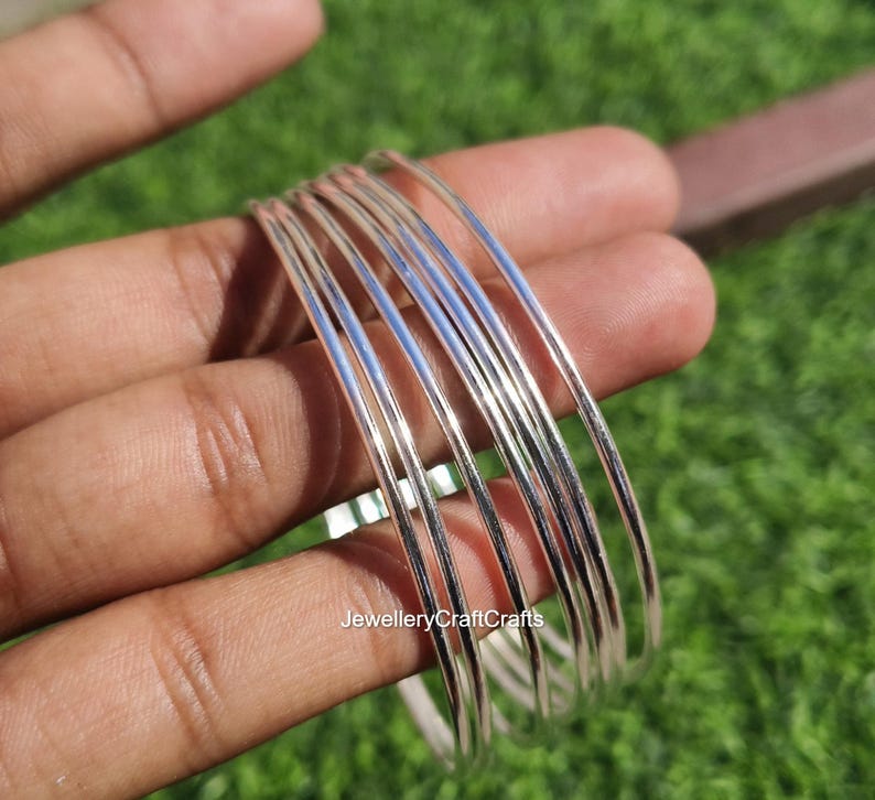 Peut inclure: Un ensemble de bracelets fins en argent. Les bracelets sont lisses et r&eacute;fl&eacute;chissants, avec un &eacute;clat m&eacute;tallique brillant. Les bijoux sont tenus dans une main sur fond d'herbe verte.
