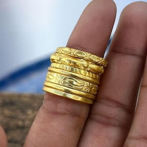 Könnte beinhalten: Ein Stapel aus sechs Goldringen an einem Finger. Die Ringe weisen verschiedene Designs auf, darunter glatte Bänder, texturierte Oberflächen mit blattartigen Mustern und gravierte geometrische Motive. Jeder Ring hat ein poliertes, gelbgoldenes Finish.