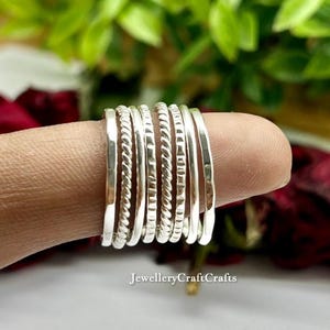Puede incluir: Un conjunto de anillos de plata apilados en un dedo. Cada anillo tiene un diseño único: lisos, trenzados o texturizados. El fondo borroso muestra hojas verdes y rojas. El conjunto es elegante y resalta las joyas.