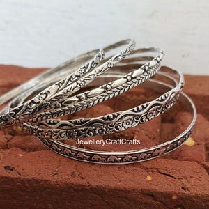 Könnte beinhalten: Eine Sammlung silberner Armreifen mit aufwendigen Blumen- und Blattmustern. Die Armreifen liegen auf einer strukturierten, rotbraunen Oberfläche. Der Text "JewelleryCraftCrafts" ist auf dem unteren Armreif zu sehen.