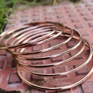 May include: A collection of rose gold bangles. The bangles are thin, circular, and have a polished surface. The jewellery is presented on a reddish-brown surface. The bangles are stacked, creating a layered effect. The image is well-lit, highlighting the metallic sheen of the bangles.