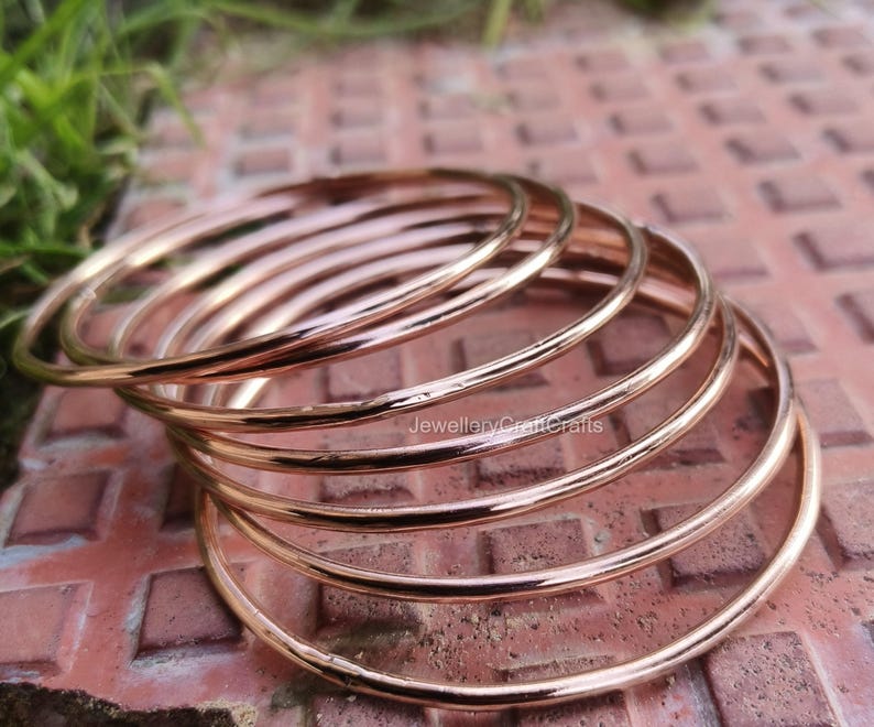 May include: A collection of rose gold bangles. The bangles are thin, circular, and have a polished surface. The jewellery is presented on a reddish-brown surface. The bangles are stacked, creating a layered effect. The image is well-lit, highlighting the metallic sheen of the bangles.