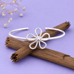 Könnte beinhalten: Ein silberfarbenes Armband mit einem zentralen, durchbrochenen Blumenmotiv mit fünf abgerundeten Blütenblättern. Das Armband hat eine glatte, polierte Oberfläche und ein kreisförmiges Band, das hinten offen ist und ein schlichtes Design aufweist.