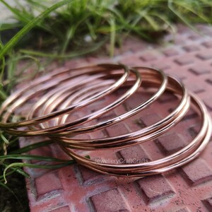 May include: A collection of thin, rose gold bangles. The bangles are smooth and shiny, reflecting light. The jewellery is displayed on a textured, reddish-brown surface with green grass in the background. The text "JewelleryCraftCrafts" is visible.