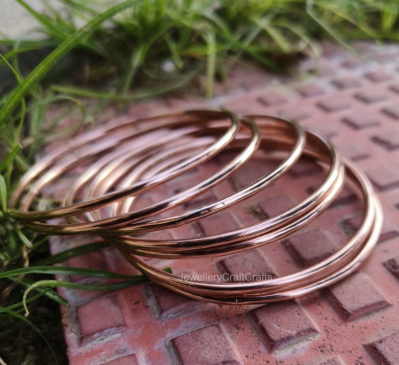 May include: A collection of thin, rose gold bangles. The bangles are smooth and shiny, reflecting light. The jewellery is displayed on a textured, reddish-brown surface with green grass in the background. The text "JewelleryCraftCrafts" is visible.