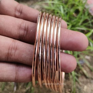 May include: A collection of rose gold bangles. The smooth, metallic bracelets are stacked together, reflecting light. The jewellery is held in a hand, with a blurred green background.