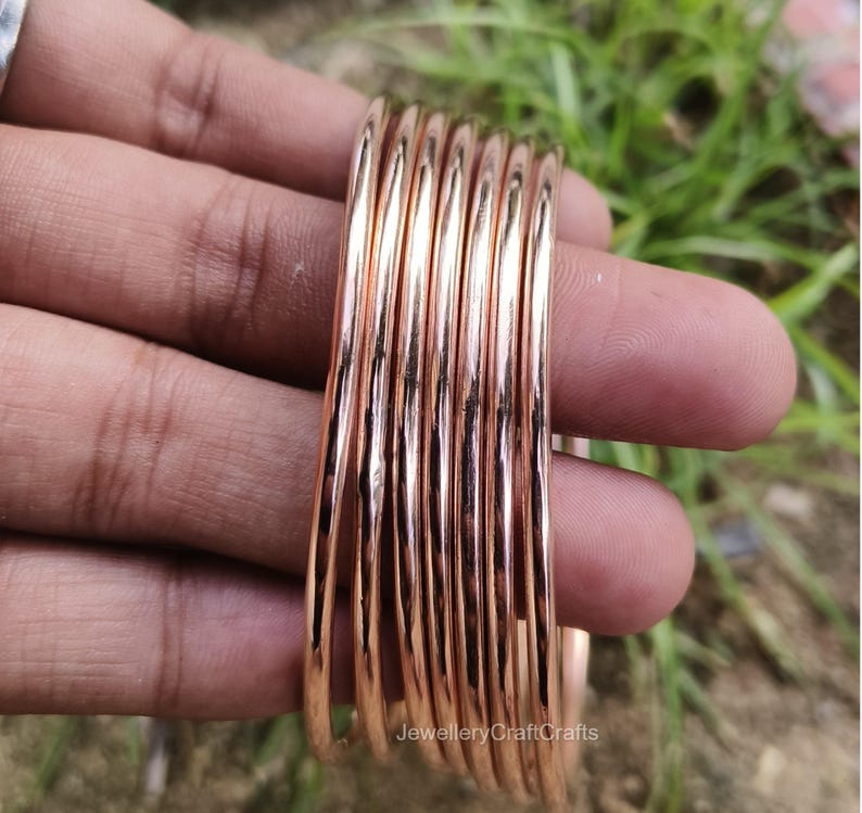 May include: A collection of rose gold bangles. The smooth, metallic bracelets are stacked together, reflecting light. The jewellery is held in a hand, with a blurred green background.