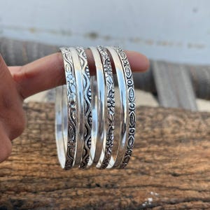 Könnte beinhalten: Ein Set aus sechs silbernen Armreifen mit aufwendigen floralen und geometrischen Mustern. Die Armreifen werden auf einer Holzoberfläche präsentiert, die ihre detaillierten Designs und ihr poliertes Finish hervorhebt. Jeder Armreif hat ein einzigartiges Muster.