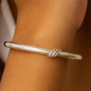 Könnte beinhalten: Ein silbernes Armreif-Armband mit gehämmertem Finish und gedrehtem Design. Das Armband ist ein schlichtes, elegantes Schmuckstück. Das Armband wird am Arm einer Person gezeigt.