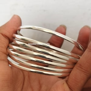 Könnte beinhalten: Ein Stapel silberner Armreifen. Die Armreifen sind glatt, glänzend und rund und reflektieren das Licht. Der Schmuck wird vor einem neutralen Hintergrund präsentiert. Die Armreifen sind ein klassisches Accessoire.
