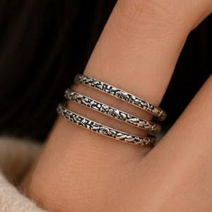 Könnte beinhalten: Drei silberne Ringe mit einem detaillierten, strukturierten Muster sind auf einem Finger gestapelt. Die Ringe haben ein Vintage- oder Antik-Aussehen mit einem dunklen, oxidierten Finish, das das komplizierte Design hervorhebt. Die Ringe sind ein Schmuckstück.