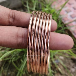 May include: A collection of rose gold bangles. The bangles are slender, circular, and have a shiny surface. The bangles are held in a hand against a backdrop of green grass and a blurred surface.