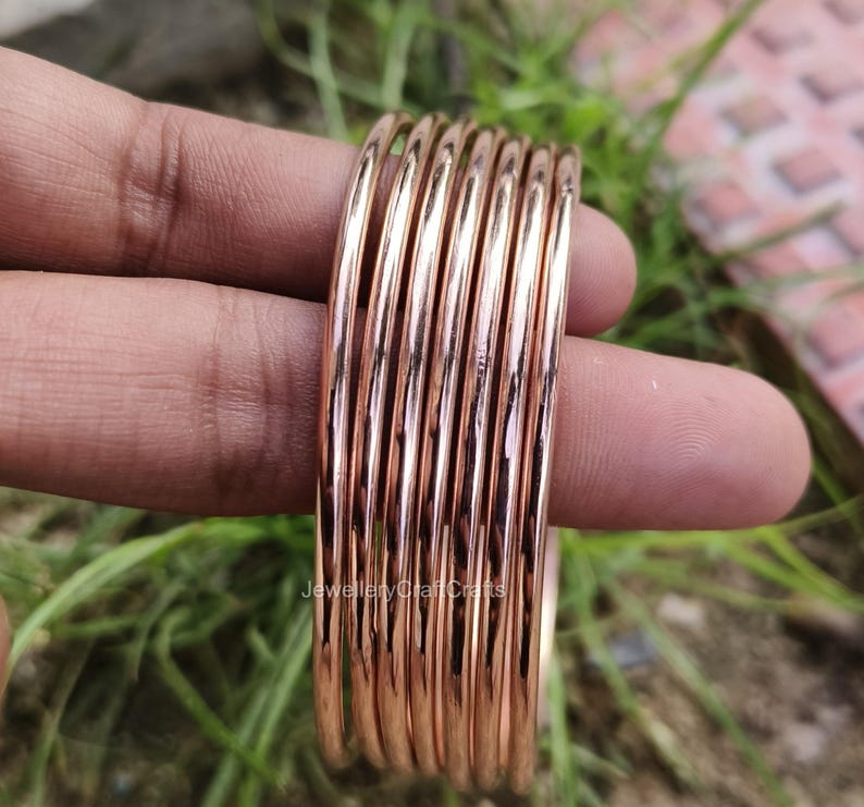 May include: A collection of rose gold bangles. The bangles are slender, circular, and have a shiny surface. The bangles are held in a hand against a backdrop of green grass and a blurred surface.