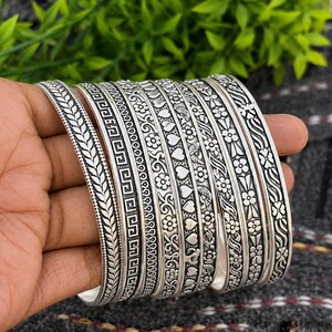 Könnte beinhalten: Eine Sammlung silberner Armreifen mit aufwendigen Designs. Die Armreifen weisen Muster wie Blätter, geometrische Formen, Blumen und Herzen auf. Die Armbänder werden auf einer Hand präsentiert, um die Vielfalt der Stile zu zeigen.