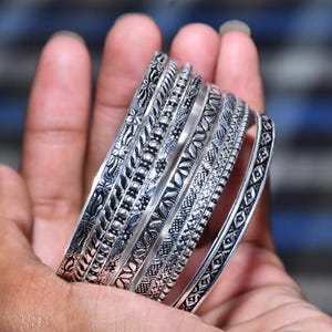 Könnte beinhalten: Ein Set aus sieben silberfarbenen Metallarmreifen mit verschiedenen geprägten Mustern, darunter florale Motive, geometrische Formen und Perlenstrukturen. Jeder Armreif hat ein oxidiertes Finish, das die eingravierten Details hervorhebt.