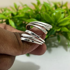 Könnte beinhalten: Ein silberfarbener Ring mit einem einzigartigen Spiraldesign. Der Ring hat mehrere gebogene Bänder, die sich um den Finger wickeln und einen offenen, modernen Look erzeugen. Der Ring wird vor einem verschwommenen grünen Hintergrund gezeigt.