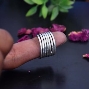 Könnte beinhalten: Ein silberfarbener Ring mit einem Multi-Band-Design, das aus sieben dünnen, glatten, parallelen, kreisförmigen Drähten besteht, die zu einem einzigen breiten Band zusammengefügt sind, das den Finger umschließt und eine polierte Oberfläche aufweist.