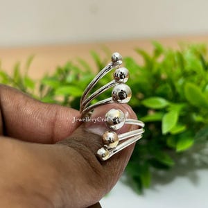 Könnte beinhalten: Ein silberfarbener Ring mit einem einzigartigen Design. Der Ring hat mehrere gebogene Bänder, die jeweils mit kleinen, kugelförmigen Akzenten verziert sind. Der Ring wird von einer Hand vor einem Hintergrund aus grünem Laub gehalten.