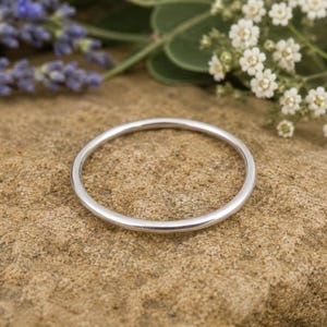 Könnte beinhalten: Ein schlichter, silberner Ring mit glatter, polierter Oberfläche. Der Ring ist kreisförmig und liegt auf einer strukturierten, beigefarbenen Oberfläche, mit Zweigen weißer Blumen und Lavendel im Hintergrund. Ein minimalistischer Schmuck.