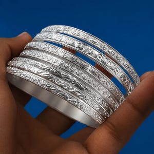 Könnte beinhalten: Ein Set aus fünf silberfarbenen Armreifen mit komplexen, geprägten Blumen- und Geometriemustern. Jeder Armreif hat eine polierte metallische Oberfläche mit detaillierten Gravuren, die sich um den gesamten Umfang der runden Bänder ziehen.