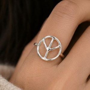 Könnte beinhalten: Ein silberner Ring mit einem Friedenssymbol-Design. Der Ring hat eine kreisförmige Form mit drei Linien, die von einem zentralen Punkt nach außen verlaufen. Der Ring wird an einem Finger getragen.