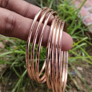 May include: A set of thin, rose gold bangles. The bangles are smooth and shiny, reflecting light. The jewellery is held in a hand against a background of green foliage.