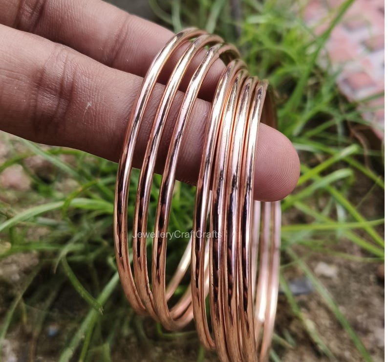 May include: A set of thin, rose gold bangles. The bangles are smooth and shiny, reflecting light. The jewellery is held in a hand against a background of green foliage.