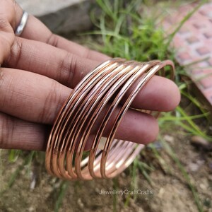 May include: A collection of copper-coloured bangles, held in a hand. The bangles are thin, round, and stacked together. The jewellery has a shiny, metallic finish. The background is blurred, with green grass and a hint of brown.