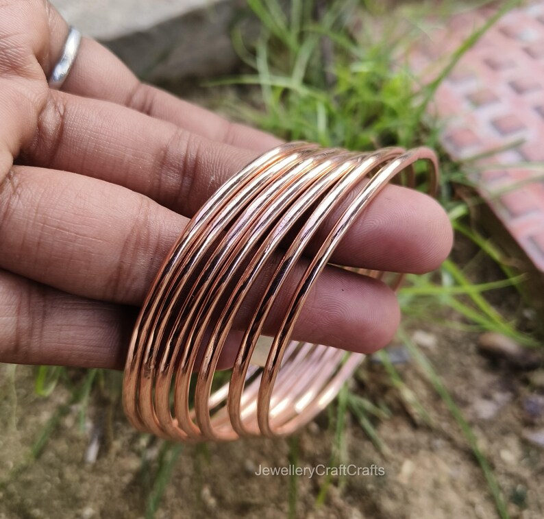 May include: A collection of copper-coloured bangles, held in a hand. The bangles are thin, round, and stacked together. The jewellery has a shiny, metallic finish. The background is blurred, with green grass and a hint of brown.