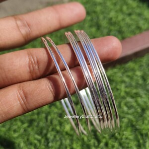 Peut inclure: Un ensemble de bracelets fins de couleur argent&eacute;e. Les bracelets sont lisses et r&eacute;fl&eacute;chissants, avec une l&eacute;g&egrave;re courbure. Les bijoux sont tenus dans une main sur un fond d'herbe verte et d'une surface marron. Le texte "JewelleryCraftCrafts" est visible.