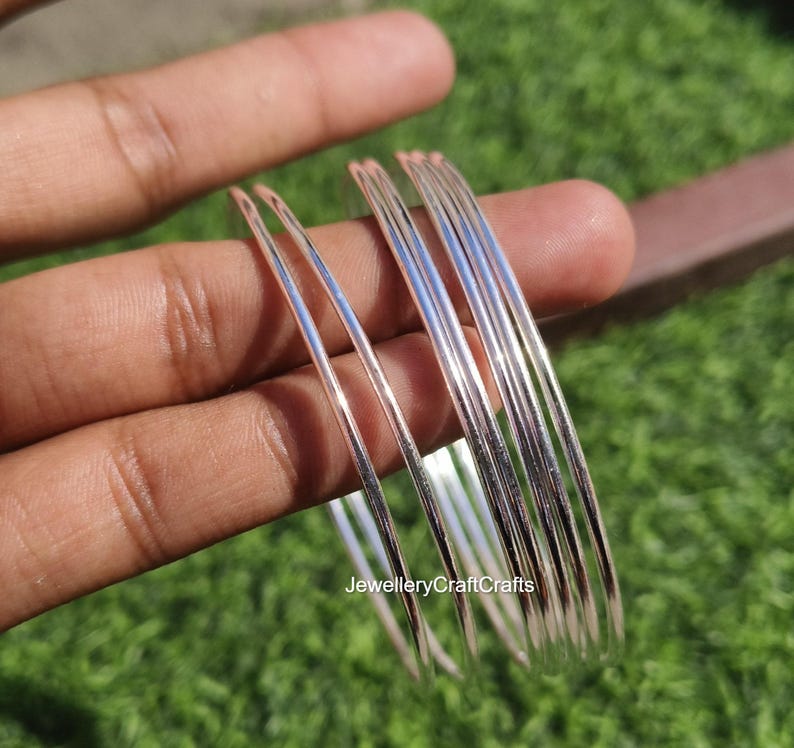 Peut inclure: Un ensemble de bracelets fins de couleur argent&eacute;e. Les bracelets sont lisses et r&eacute;fl&eacute;chissants, avec une l&eacute;g&egrave;re courbure. Les bijoux sont tenus dans une main sur un fond d'herbe verte et d'une surface marron. Le texte "JewelleryCraftCrafts" est visible.