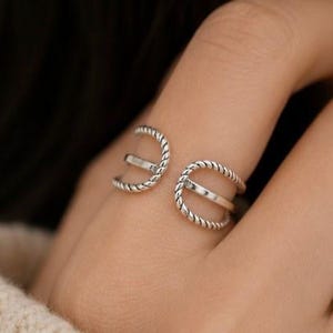 Könnte beinhalten: Ein silberner Ring mit einem einzigartigen Design. Der Ring hat zwei offene Kreise, die durch einen horizontalen Balken verbunden sind. Die Ränder der Kreise haben eine gedrehte, seilartige Textur, die dem Schmuck Detail verleiht.
