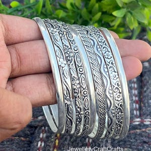 May include: A stack of silver bangles with intricate floral and geometric patterns. The bangles are held in a hand, showcasing the detailed designs. The jewelry is displayed against a natural green and gray background. Text on the image reads "JewelleryCrafts".