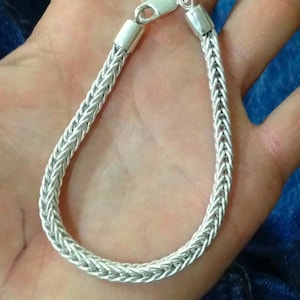 Könnte beinhalten: Ein silberfarbenes Kettenarmband mit einem geflochtenen Weizen- oder Fuchsschwanz-Gliedermuster. Das Armband ist mit glatten zylindrischen Endkappen und einem sicheren Karabinerverschluss versehen, was eine durchgehende, flexible Metallschleife bildet.