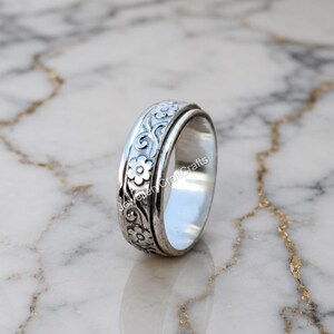 Könnte beinhalten: Ein silberner Ring mit floralem Design. Der Ring hat ein Band mit geprägten Blumen und Blättern. Der Ring wird auf einer Marmoroberfläche gezeigt. Der Ring ist etwa 1,9 cm breit.