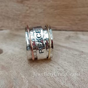 Könnte beinhalten: Ein silberfarbener Spinner-Ring mit dem Wort "PEACE" auf dem Mittelband eingraviert. Der Ring hat eine strukturierte Oberfläche und verfügt über Bänder in Roségold- und Goldtönen. Der Ring wird auf einer Holzoberfläche präsentiert.