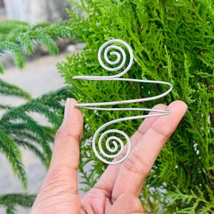 Könnte beinhalten: Ein Oberarmreif aus silberfarbenem Metall mit einem durchgehenden Drahtdesign, das an beiden Enden zwei markante Spiralen bildet. Die Struktur besteht aus einem einzigen Stück poliertem Draht, das zu einer Reihe offener Schlaufen geformt ist.