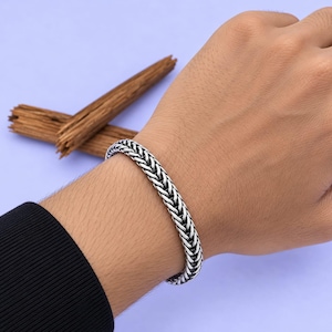 Könnte beinhalten: Ein silberfarbenes Armband mit einem geflochtenen Weizenketten-Design. Die Metallglieder sind eng miteinander verbunden und erzeugen ein strukturiertes, abgerundetes Erscheinungsbild mit dunklen, oxidierten Details zwischen den Gliedern.