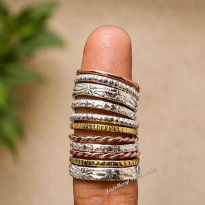 May include: A collection of stackable rings in various designs and metals, including silver, gold, and copper. The rings are worn on a finger, showcasing their different textures and patterns. The rings have etched and twisted designs.