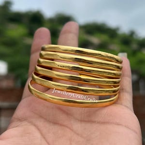 Könnte beinhalten: Ein Stapel goldfarbener Armreifen, jeder mit einem glatten, abgerundeten Design. Die Armreifen sind übereinander gestapelt, wodurch ein geschichteter Effekt entsteht. Der Text "JewelleryCraftCrafts" ist sichtbar.