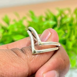 Op de afbeelding: Een zilverkleurige ring met een dubbel V-vormig ontwerp. De ring wordt in een hand gehouden, met een vage groene achtergrond. De ring heeft een minimalistische stijl en is geschikt voor dagelijks gebruik.