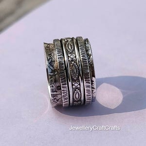 Könnte beinhalten: Ein silberner Spinner-Ring mit mehreren Bändern, die jeweils aufwendige geprägte Muster aufweisen. Die Bänder des Rings weisen florale und geometrische Designs auf, wodurch ein strukturiertes und detailliertes Aussehen entsteht. Der Ring wird auf einer hellen Oberfläche präsentiert.