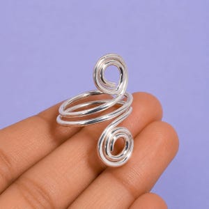 Könnte beinhalten: Ein silberfarbener Ring mit einem Bypass-Design und zwei offenen, spiralförmigen Enden. Das Band besteht aus einem durchgehenden Draht, der sich zur Kreisform windet, wobei jedes Ende in einer ausgeprägten, gewickelten Spirale ausläuft.