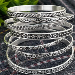 Könnte beinhalten: Ein Stapel silberner Armreifen mit aufwendigen floralen und geflochtenen Designs. Die Armreifen variieren in der Breite und im Muster und zeigen detaillierte Handwerkskunst. Der Schmuck wird auf einer strukturierten, dunkelgrauen Oberfläche präsentiert.