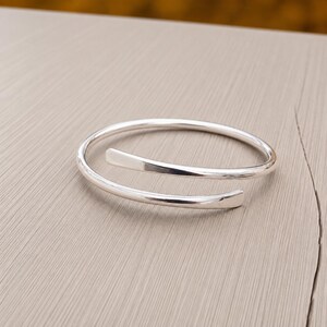 Könnte beinhalten: Ein poliertes silberfarbenes Bypass-Armband mit einem glatten, durchgehenden Metallband, das sich zu einem offenen, überlappenden Design windet. Die Enden des Armbands sind leicht abgeflacht und verjüngt und weisen eine reflektierende Oberfläche auf.