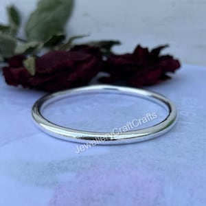 Peut inclure: Une bague argentée brillante avec un design lisse et arrondi. La bague est circulaire et semble être en métal. Le fond comprend des fleurs rouges floues et le texte "JewelleryCraftCrafts".