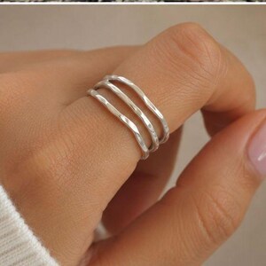 Könnte beinhalten: Ein silberner Ring mit drei Bändern wird an einem Finger getragen. Der Ring hat ein strukturiertes, gehämmertes Aussehen. Die Hand trägt einen weißen Pullover. Der Hintergrund ist eine weiche, neutrale Farbe.